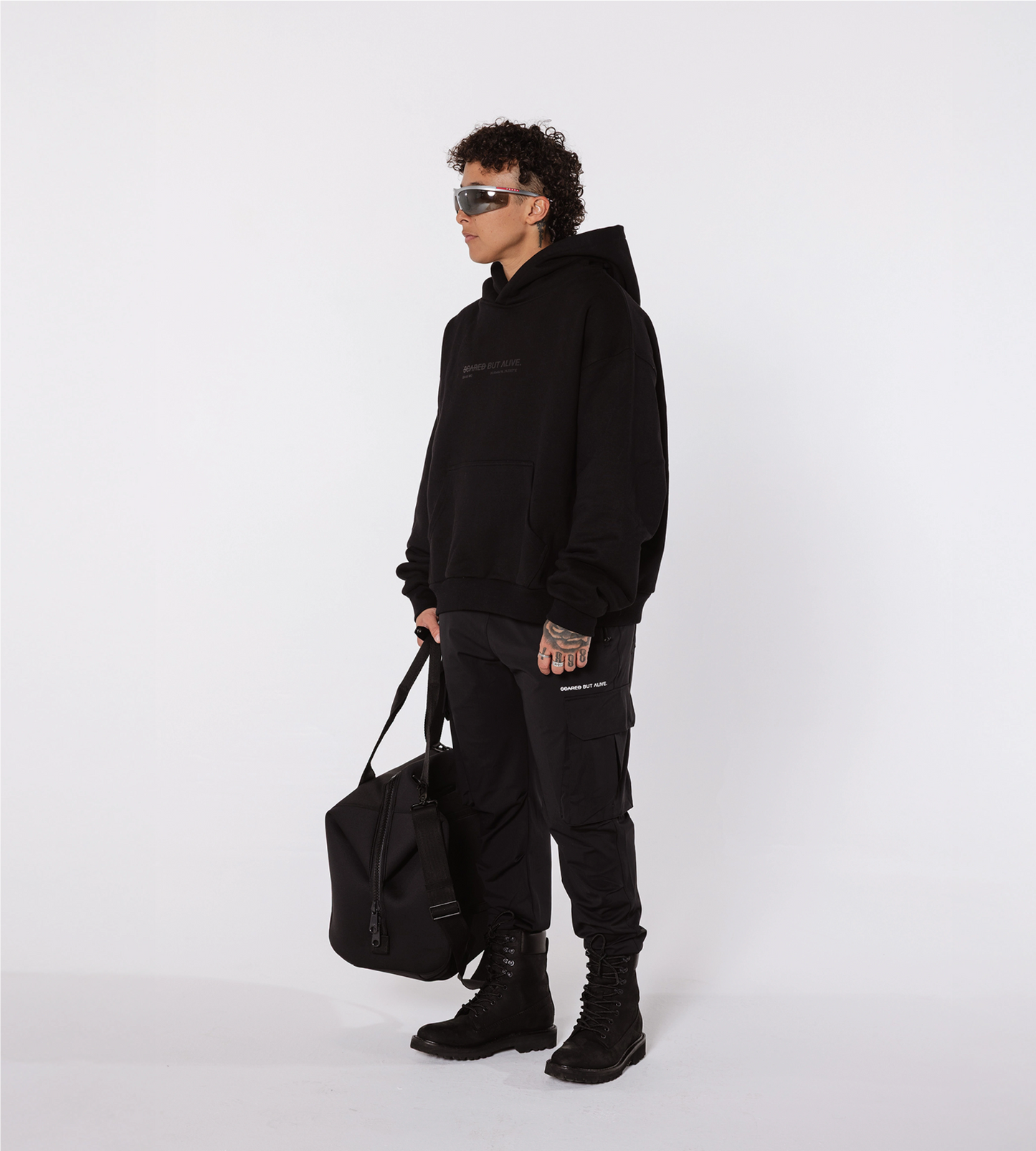 [Color: Black Out] A model in a white studio, wearing the black Los Angeles hoodie, cargo joggers, combat boots and sunglasses. The model is also holding a black duffel bag, in their right hand.