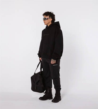 [Color: Black Out] A model in a white studio, wearing the black Los Angeles hoodie, cargo joggers, combat boots and sunglasses. The model is also holding a black duffel bag, in their right hand.