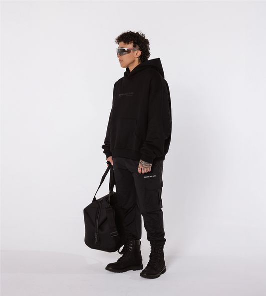 [Color: Black Out] A model in a white studio, wearing the black Los Angeles hoodie, cargo joggers, combat boots and sunglasses. The model is also holding a black duffel bag, in their right hand.