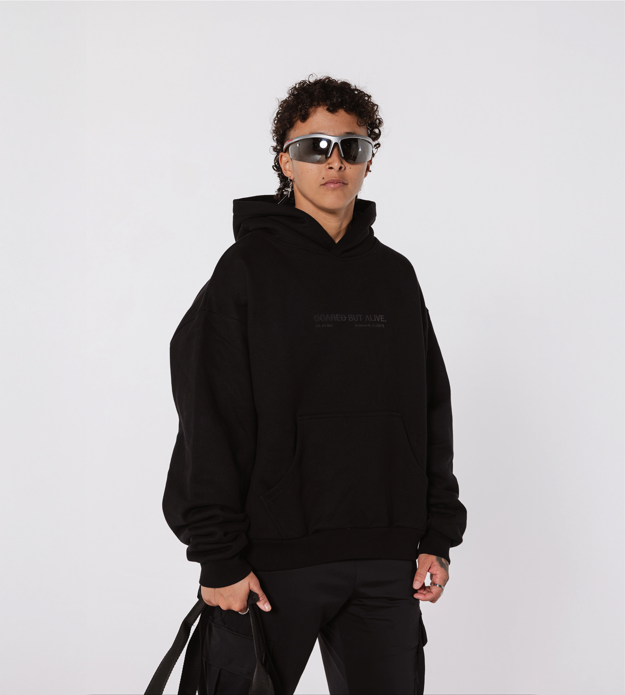 [Color: Black Out] Model wearing the LA Hodie with Scared But Alive text detail on the front, paired with black cargo pants and futuristic wraparound sunglasses.