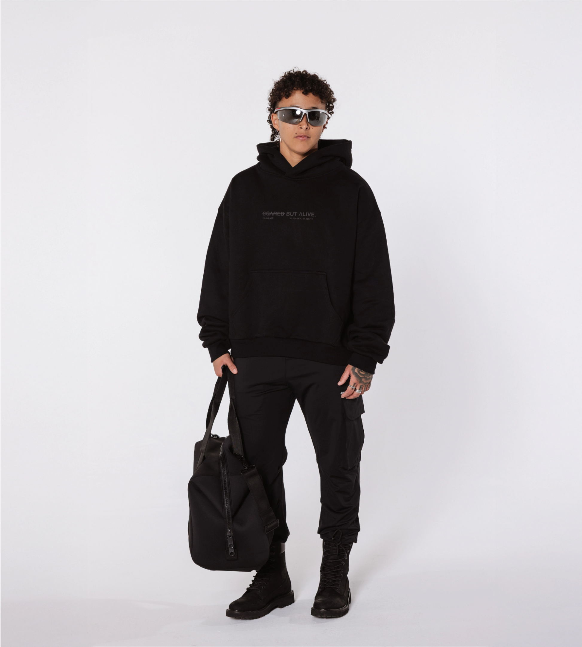 [Color: Black Out] Model wearing sunglasses, the black Los Angeles hoodie, cargo joggers, and combat boots. The model is also carrying a black duffel bag in their right hand.