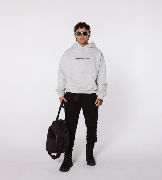 [Color: Grey Matter] Full shot of a model dressed in the Los Angeles gray hoodie, black cargo joggers, and boots. The model is holding a duffel bag.